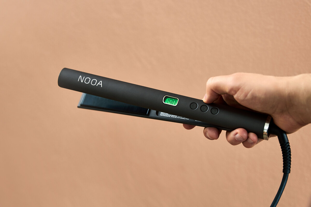 A close-up picture of a dark grey NOOA hair straightener in a person's hand with a peach colored background