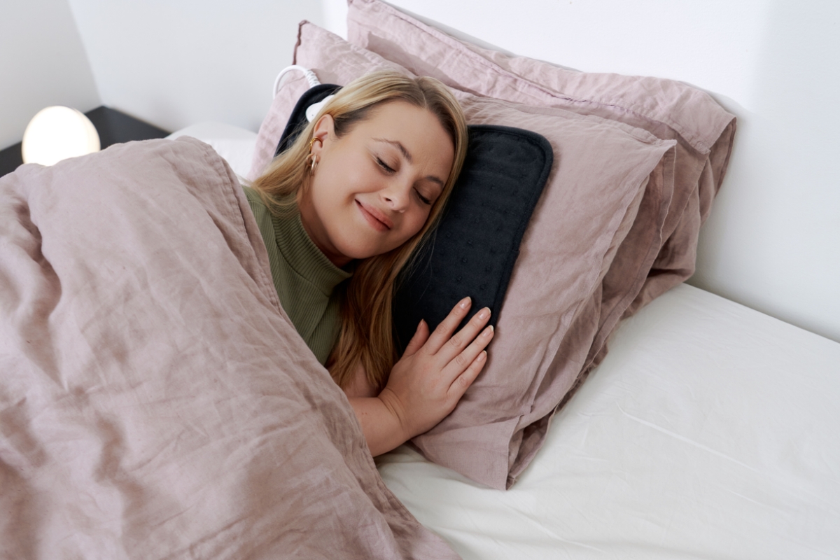 A smiling woman with light-colored hair laying on a bed with pink sheets and laying her head on a black Point heating pad