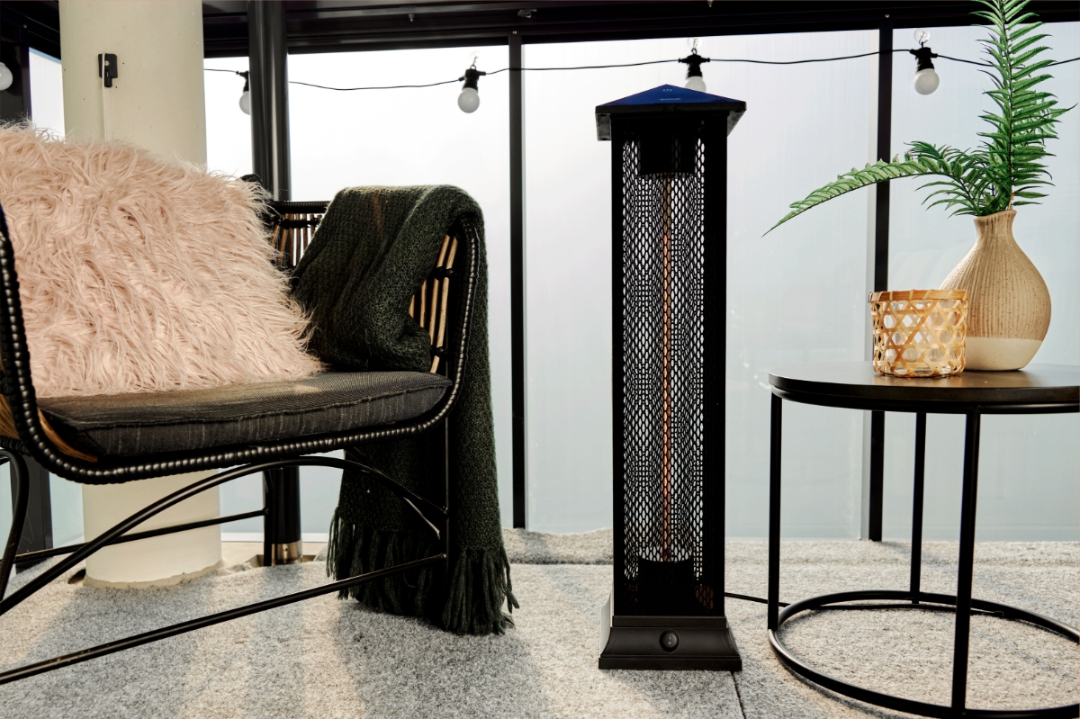 A wide image of the POINT PRO POPHTOW81 patio heater on a balcony. On the left to it is a lounge chair with pink throw pillow and a green blanket. On the right is a coffee table with a plant and a woven candle holder on it.